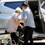 Caregiver helping disabled elderly woman in wheelchair to get into the car,helpful daughter care and support senior mother to stand up from wheelchair in outdoor, caring for old people