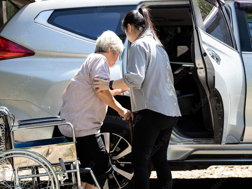 Caregiver helping disabled elderly woman in wheelchair to get into the car,helpful daughter care and support senior mother to stand up from wheelchair in outdoor, caring for old people