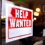 Sign text closeup for help wanted with red and white colors by entrance to store shop business building during corona virus covid 19 pandemic