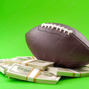 Winning bet on sporting event, money in sport and sports betting conceptual idea with american football ball and wads of cash isolated on green background