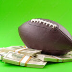 Winning bet on sporting event, money in sport and sports betting conceptual idea with american football ball and wads of cash isolated on green background