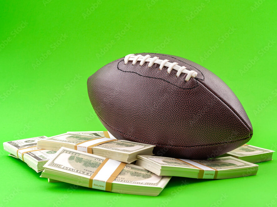 Winning bet on sporting event, money in sport and sports betting conceptual idea with american football ball and wads of cash isolated on green background
