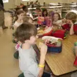 This image depicts a group of school children, who were seated in the lunchroom of a metropolitan Atlanta, Georgia primary school taking their daily lunch break during their school day activities. In this particular view, seated in the foreground, were two playful boys, one of whom was about to begin eating his whole-wheat sandwich, minus the crust. Hopefully, his lunch included some fresh fruit, as was the case for some of his classmates