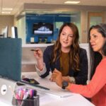 Two business women talking about sales in office at desk with laptop